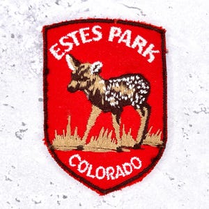May include: A shield-shaped embroidered patch with a bright red background. It features a brown and white spotted fawn standing in tan grass. The text ESTES PARK is arched at the top and COLORADO is at the bottom, both in white lettering.