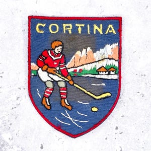 May include: A shield-shaped embroidered patch with a blue background and a red border. It features the yellow text CORTINA above an ice hockey player in a red jersey and white shorts, holding a yellow stick on ice with mountains in the back.