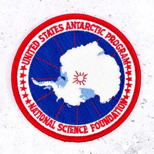 Può includere: Toppa rotonda ricamata con il logo del Programma Antartico degli Stati Uniti. La toppa ha un bordo rosso con testo e stelle bianche. Il centro mostra una mappa bianca dell'Antartide su sfondo blu, con un sole rosso.