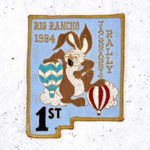 Könnte beinhalten: Gestickter Aufnäher mit hellblauem Hintergrund, der einen braunen Jackrabbit zeigt, der zwei Heißluftballons hält. Der Text enthält "Rio Rancho 1984", "Jackrabbit Rally" und "1st". Der Aufnäher hat einen goldenen Rand.