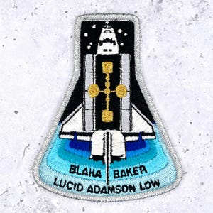 May include: Embroidered patch featuring a stylized space shuttle design. The patch has a black and white body with gold accents and a blue and turquoise gradient base. The text "BLAHA BAKER LUCID ADAMSON LOW" is embroidered on the bottom.