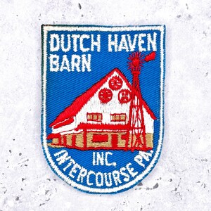 May include: Embroidered patch depicting a barn with a red roof and windmill on a blue backdrop. The text "DUTCH HAVEN BARN" and "INTERCOURSE PA. INC." are in white and blue. The patch is edged with a white border.