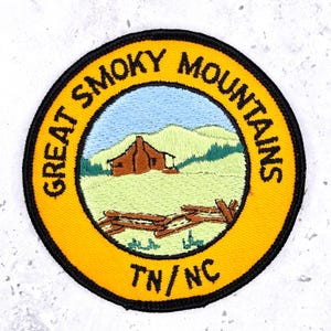 Op de afbeelding: Ronde geborduurde patch met een gele rand en zwarte omtrek. De patch toont de tekst "GREAT SMOKY MOUNTAINS". Binnenin is een landschap met een hut, bergen en de tekst "TN/NC".