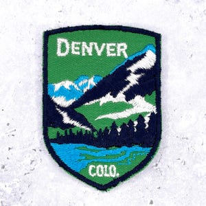 May include: Embroidered patch featuring a mountain landscape with the word "DENVER" at the top and "COLO." at the bottom. The patch has a green background with blue, white, and dark blue mountain and water details.