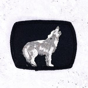 May include: Embroidered patch featuring a howling wolf in shades of gray and white against a black background. The patch has a rectangular shape with rounded corners and a stitched border. Ideal for jackets, bags, or other fabric items.