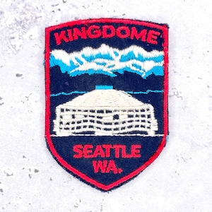 Puede incluir: Un parche bordado en forma de escudo con fondo azul oscuro y borde rojo. Presenta el texto blanco KINGDOME en la parte superior, una cordillera blanca y azul claro, un estadio con cúpula blanco y el texto rojo SEATTLE WA. en la parte inferior.