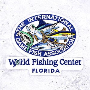 May include: Embroidered patch with a circular design showing several fish, such as a marlin and shark, in blue and white. The text reads "The International Game Fish Association" and "World Fishing Center FLORIDA."
