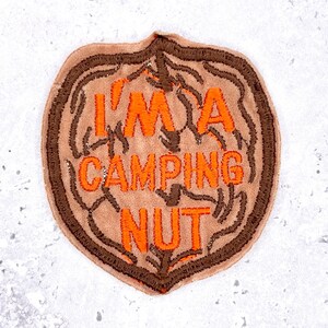 May include: A tan patch with the text "I'M A CAMPING NUT" in orange. The patch has a brown border and a design that resembles a nut. The patch is likely for a backpack or jacket.