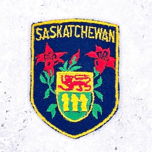 May include: Embroidered patch with the word "SASKATCHEWAN" in yellow letters. The patch features a shield design with red flowers, a coat of arms, and a yellow border on a navy blue background.