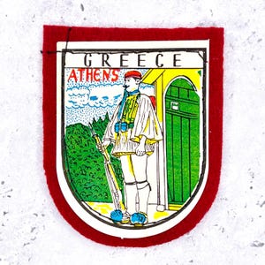Puede incluir: Un parche rectangular de fieltro rojo con una ilustración impresa de un guardia Evzone griego con uniforme tradicional sosteniendo un rifle. El texto GREECE está impreso en la parte superior, con ATHENS en rojo debajo. El guardia está junto a una puerta verde.