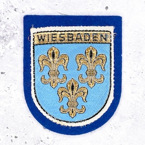 Könnte beinhalten: Ein schildförmiger, bestickter Aufnäher mit einem dunkelblauen Filzrand. Die Mitte zeigt ein hellblaues Feld mit drei goldenen Lilien. Über den Symbolen befindet sich ein goldenes Banner mit dem schwarzen Text WIESBADEN in Großbuchstaben.