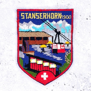 May include: Embroidered patch with a shield shape, featuring the text "STANSERHORN 1900" in yellow on a blue background. The design includes a mountain scene, a building, and a Swiss flag.
