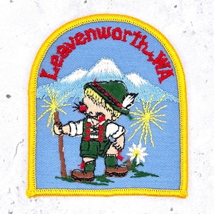 May include: An embroidered patch with a yellow border on a light blue background. It features a figure in green traditional clothing, a hat with a feather, and a walking stick. Behind the figure are mountains, yellow sunbursts, and a white flower. The text reads Leavenworth, WA.
