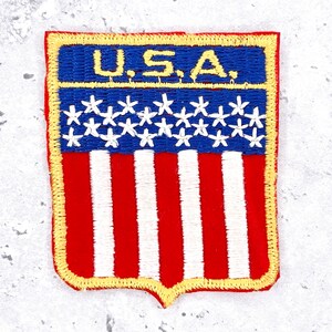 May include: A rectangular embroidered patch featuring the text U.S.A. in yellow thread on a blue background. Below the text is a blue section with white embroidered stars, followed by a red and white vertical striped section at the bottom.