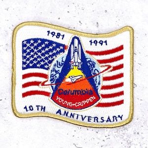 Pode incluir: Um patch retangular com uma borda dourada apresenta um design que comemora o 10º aniversário da missão Columbia Young-Crippen. O patch inclui uma bandeira americana, um ônibus espacial e os anos de 1981 e 1991. O texto "Columbia Young-Crippen" também é visível.