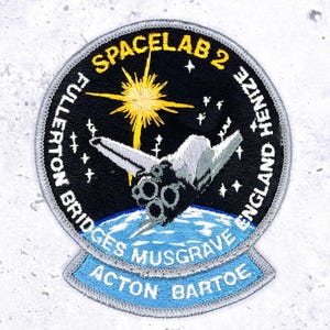 May include: Embroidered patch featuring a space shuttle design with the text "SPACELAB 2" and names. The patch has a black background with a yellow starburst and a blue and white Earth. The patch also includes the names of the crew.