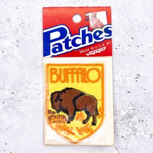 Pode incluir: Um patch vintage "Buffalo" em um cartão vermelho e azul. O patch apresenta um bisonte marrom bordado em um fundo amarelo com a palavra "BUFFALO" em laranja. O cartão diz "Patches" e "MADE IN U.S.A. BY Voyager".