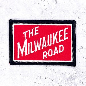 May include: Embroidered rectangular patch featuring the text "THE MILWAUKEE ROAD" in white on a red background. The patch has a black border and a white border. A vintage-style item.
