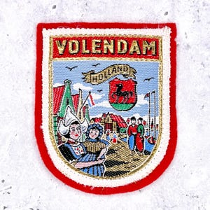 May include: A rectangular embroidered patch with a red border featuring the text VOLENDAM in gold letters. The design depicts a scene with two figures in traditional Dutch clothing, a shield with a black horse, and a banner reading HOLLAND.