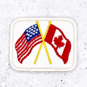 May include: A rectangular embroidered patch with a white background and a stitched border. The design features two crossed yellow flagpoles holding the flags of the United States and Canada, rendered in red, white, and blue thread colors.