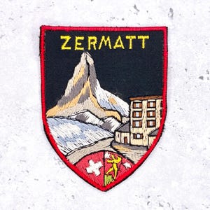 Può includere: Toppa ricamata con la scritta ZERMATT in giallo. Il design presenta un paesaggio montano, un edificio e uno scudo con una croce e una figura. La toppa ha uno sfondo nero e un bordo rosso.
