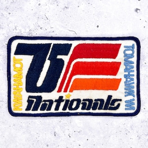 Puede incluir: Parche bordado con borde azul marino y fondo blanco. El parche presenta el texto "75 Nationals" en azul marino, con "KWAMHAMOT" y "TOMAHAWK" en amarillo y azul. El diseño incluye rayas rojas, naranjas y blancas.