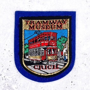 May include: A blue patch with a gold border and a detailed image of a red double-decker tram. The text "TRAMWAY MUSEUM CRICH" is displayed in gold lettering. The patch is likely a souvenir or collectible item.