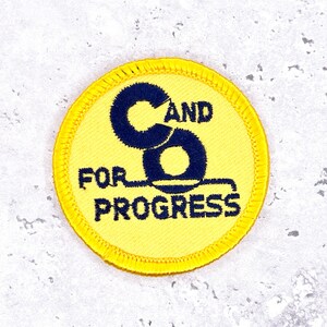 May include: A circular, yellow embroidered patch featuring the text "C and S FOR PROGRESS" in navy blue. The patch has a navy blue stylized "C" and "S" design.