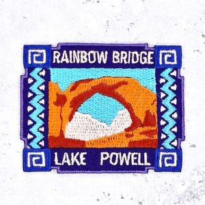 Pode incluir: Patch bordado com Rainbow Bridge e Lake Powell. O patch tem formato retangular com borda azul e o texto "RAINBOW BRIDGE" e "LAKE POWELL". A imagem mostra um arco laranja sobre um céu azul e montanhas brancas.