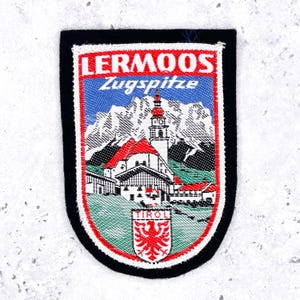 May include: A rectangular patch with a black border features a scenic illustration. The image includes a church with a red roof, mountains, and the text "LERMOOS Zugspitze." The bottom has a red and white emblem with the word "TIROL."