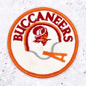 May include: Embroidered patch featuring the Tampa Bay Buccaneers logo. The circular patch has a white background with the team name in red around the edge. The logo includes a pirate helmet in red, orange, and gray.