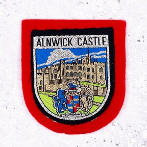 May include: A rectangular patch with a red border and a black inner border. The patch features the words "ALNWICK CASTLE" above a detailed illustration of a castle with a blue sky background. The bottom of the patch has a crest with two animals.