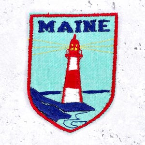 Pode incluir: Patch bordado com um design de farol. O farol tem listras vermelhas e brancas, um topo vermelho e a palavra "MAINE" em azul. Definido contra um fundo azul claro, com ondas azuis escuras e uma borda vermelha.