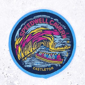 Op de afbeelding: Ronde patch met een blauwe rand en een donkerblauwe achtergrond. De patch bevat de tekst "SPEEDWELL CAVERN CASTLETON" in roze en wit. De afbeelding toont een boot met passagiers in een grot met kleurrijke rotsformaties.