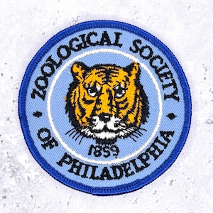 Może przedstawiać: Okrągła, haftowana naszywka z głową tygrysa w kolorze pomarańczowym i czarnym, na jasnoniebieskim tle. Tekst "ZOOLOGICAL SOCIETY OF PHILADELPHIA 1859" jest ułożony w okrąg, z ciemnoniebieską obwódką.