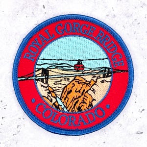 May include: A circular embroidered patch featuring a landscape of the Royal Gorge Bridge in Colorado. The design includes a red border with blue text reading Royal Gorge Bridge and Colorado, a light blue sky, and a red aerial tram car.