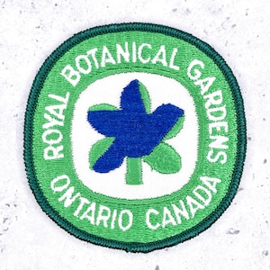 May include: A circular embroidered patch with a dark green border. The outer ring is light green with the white text ROYAL BOTANICAL GARDENS ONTARIO CANADA. The centre is white with a blue stylised bird shape over a green leaf design.