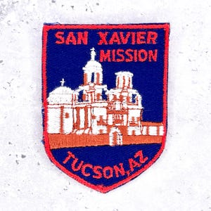 May include: A shield-shaped embroidered patch with a dark blue background and a red border. It features a white stitched illustration of the San Xavier Mission building. The text SAN XAVIER MISSION is stitched in red at the top and TUCSON, AZ at the bottom.