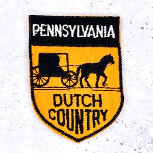 May include: Embroidered patch with a shield shape, featuring the text "PENNSYLVANIA" above a black silhouette of a horse-drawn carriage and the text "DUTCH COUNTRY" on a yellow background.