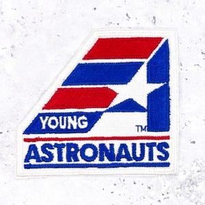 May include: Embroidered patch with a white background, featuring a red and blue geometric design and a white star. The text "YOUNG ASTRONAUTS" is embroidered in blue, with a trademark symbol. The patch has a rectangular shape with a diagonal cut.