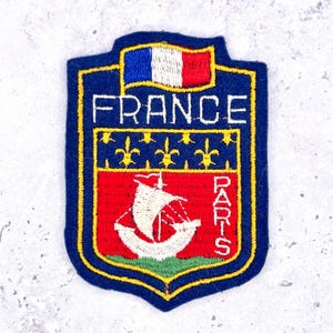 May include: Embroidered patch featuring the French flag and the words "FRANCE" and "PARIS". The patch has a blue border and a red and white sailboat design. Gold fleur-de-lis symbols are also present.
