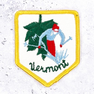 May include: A shield-shaped embroidered patch with a white background and a yellow border. It features a skier in a red hat and red pants, a light blue shirt, and white skis, positioned in front of a large green maple leaf. The text Vermont is embroidered in green below.