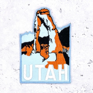 Op de afbeelding: Geborduurde patch in de vorm van Utah, met een berglandschap in oranje, zwart en wit. De omtrek van de staat is lichtblauw, met het woord "UTAH" in witte blokletters onderaan.