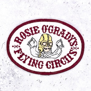 May include: An oval embroidered patch with a white background and a dark red border. The center features a yellow aviator cap with goggles and a scarf. The text "ROSIE O'GRADY'S FLYING CIRCUS" is embroidered in dark red lettering around the central figure.