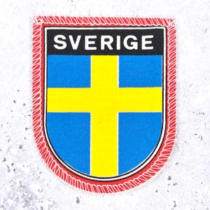 Peut inclure: Un écusson brodé en forme de bouclier représentant le drapeau suédois avec une croix jaune sur fond bleu. Le mot SVERIGE est imprimé en lettres capitales blanches sur une bande noire horizontale en haut, bordé d'un contour rouge cousu.