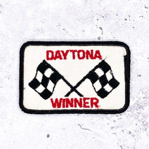 May include: Rectangular patch with the words "DAYTONA WINNER" in red letters. Two crossed checkered flags are in the center. The patch has a white background and a black border.