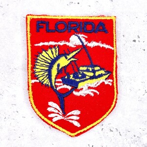 Może przedstawiać: Czerwona, haftowana naszywka z żółtą obwódką, z napisem "FLORIDA" w kolorze niebieskim. Naszywka przedstawia żółto-niebieskiego żaglicę wyskakującą z wody, z białym kwiatowym wzorem u dołu.