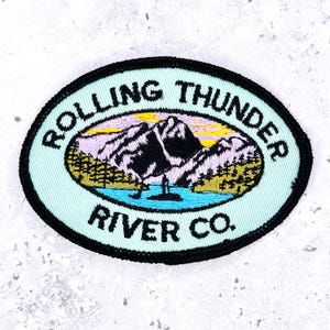May include: An oval embroidered patch with a light blue background and a black border. The design features a mountain landscape with a lake, trees, and a yellow sky. The text "ROLLING THUNDER RIVER CO." is embroidered in black around the center.