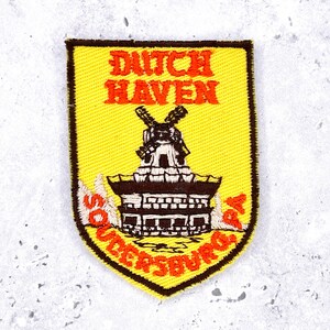 May include: A yellow embroidered patch with a shield shape. The patch features the words "DUTCH HAVEN" in orange, above a windmill illustration. The text "SOUTH HOLLAND, PA" is arched along the bottom edge.