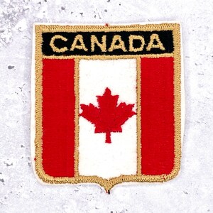 Op de afbeelding: Een rechthoekige patch met het Canadese vlagontwerp. De bovenste sectie heeft het woord "CANADA" in gouden letters op een zwarte achtergrond. Daaronder toont de vlag een rode esdoornblad op een witte achtergrond, geflankeerd door rode verticale strepen, allemaal omlijnd in goud.